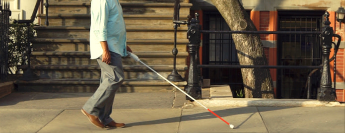 On a blue font, there is a visual featuring the functional parts of WeWALK smart cane. The handle tip of the cane is narrowing towards the top. The middle of the handle is much wider. At the bottom there is Microphone, Then in order there is Wrist Strap Hole, Soft-Touch Grip, which is a grey surface, Vibration Buttons which have a bump on both the right and left sides Touchpad, which is a white, rectangular surface Ultrasonic sensor located in a circular hole, Front Led as a small dot in the front, At the tip of WeWALK which attaches to the white cane there is White Cane Adaptor There is a regular white cane which WeWALK attaches to. At the back side of WeWALK there is a speaker in the middle