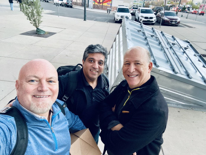Image contains a photo of three men smiling at the camera, and one of them is holding a brown cardboard box.