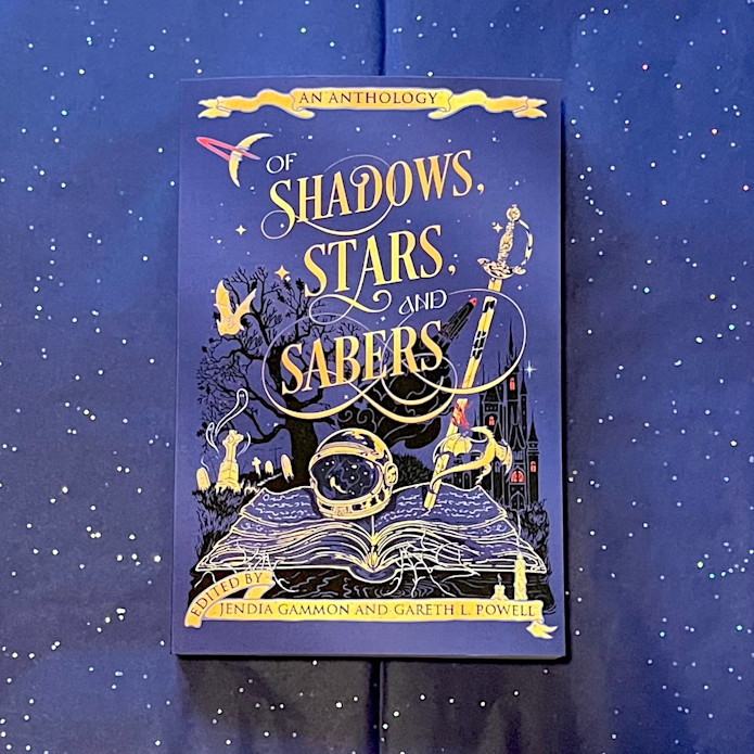 Copy of the anthology OF SHADOWS, STARS, AND SABERS edited by Jendia Gammon and Gareth L. Powell, a dark blue book with gold letters and designs against dark blue, glittery tissue paper.