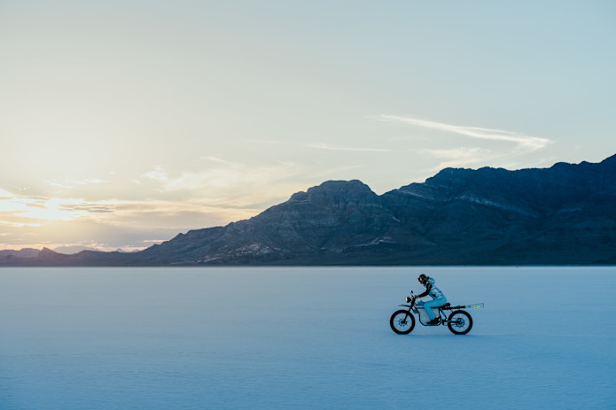  Lindsay Vonn pedals a Terra Bike Terra Prime across the vast, flat salt desert at sunrise, mountains silhouetted in the background under a partly cloudy sky.